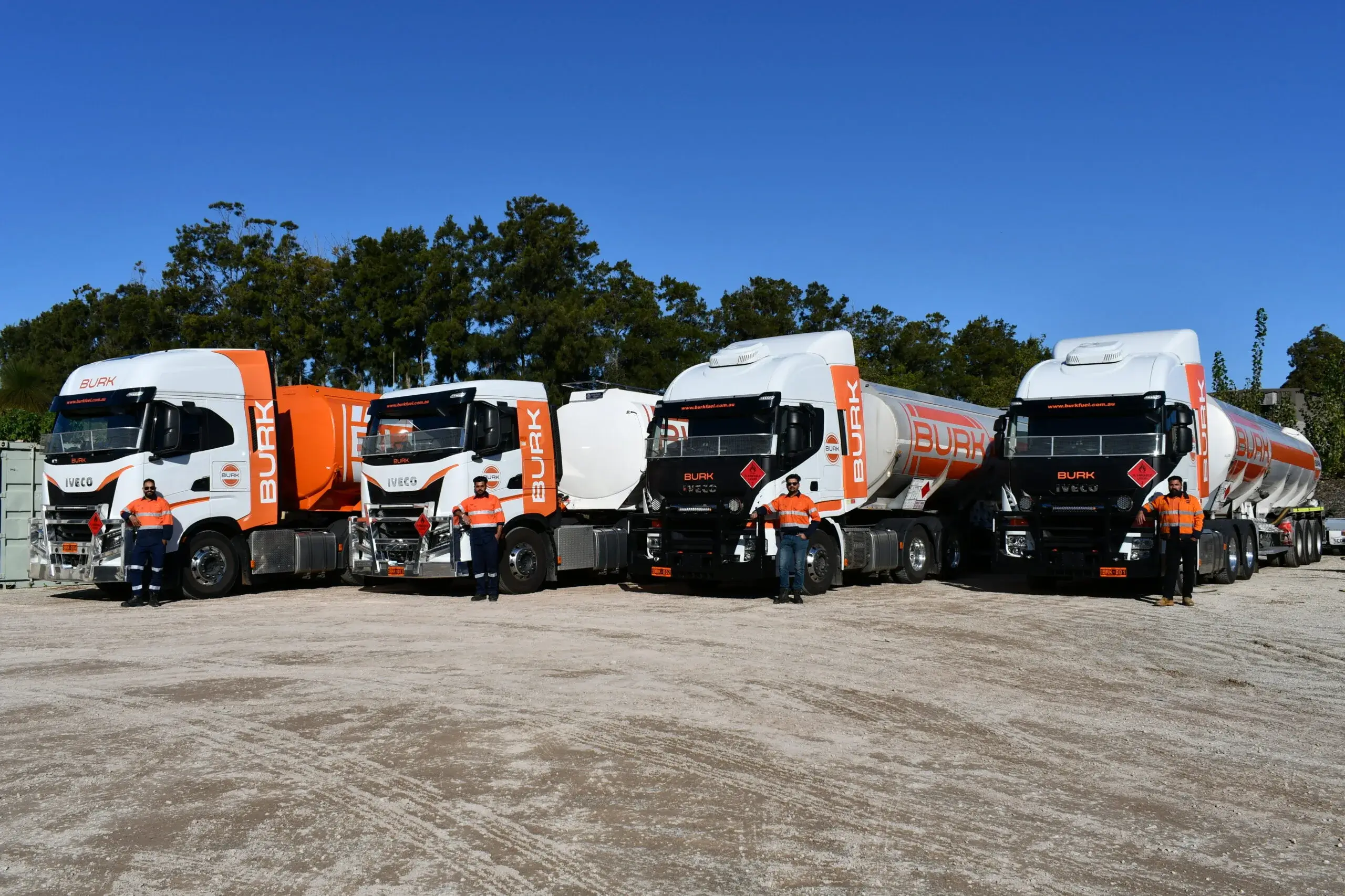 Four Burk Fuel trucks with drivers standing in front, parked under a clear blue sky.