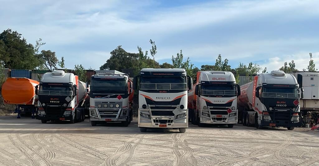 Five Burk Fuel trucks lined up in a yard with trees and containers in the background.